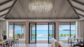 Una stanza spaziosa con una grande finestra vista spiaggia e piscina, con un lampadario moderno e un tavolo da pranzo in legno con sedie.