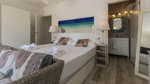 A hotel room with a bed, a wicker chair, a painting of a beach, a bathroom with a mirror, and a towel rack. - Villa Maria by Mallorca North Villas (Alcudia)