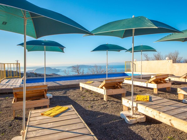 A poolside area with wooden loungers and green umbrellas, overlooking a body of water and distant hills.