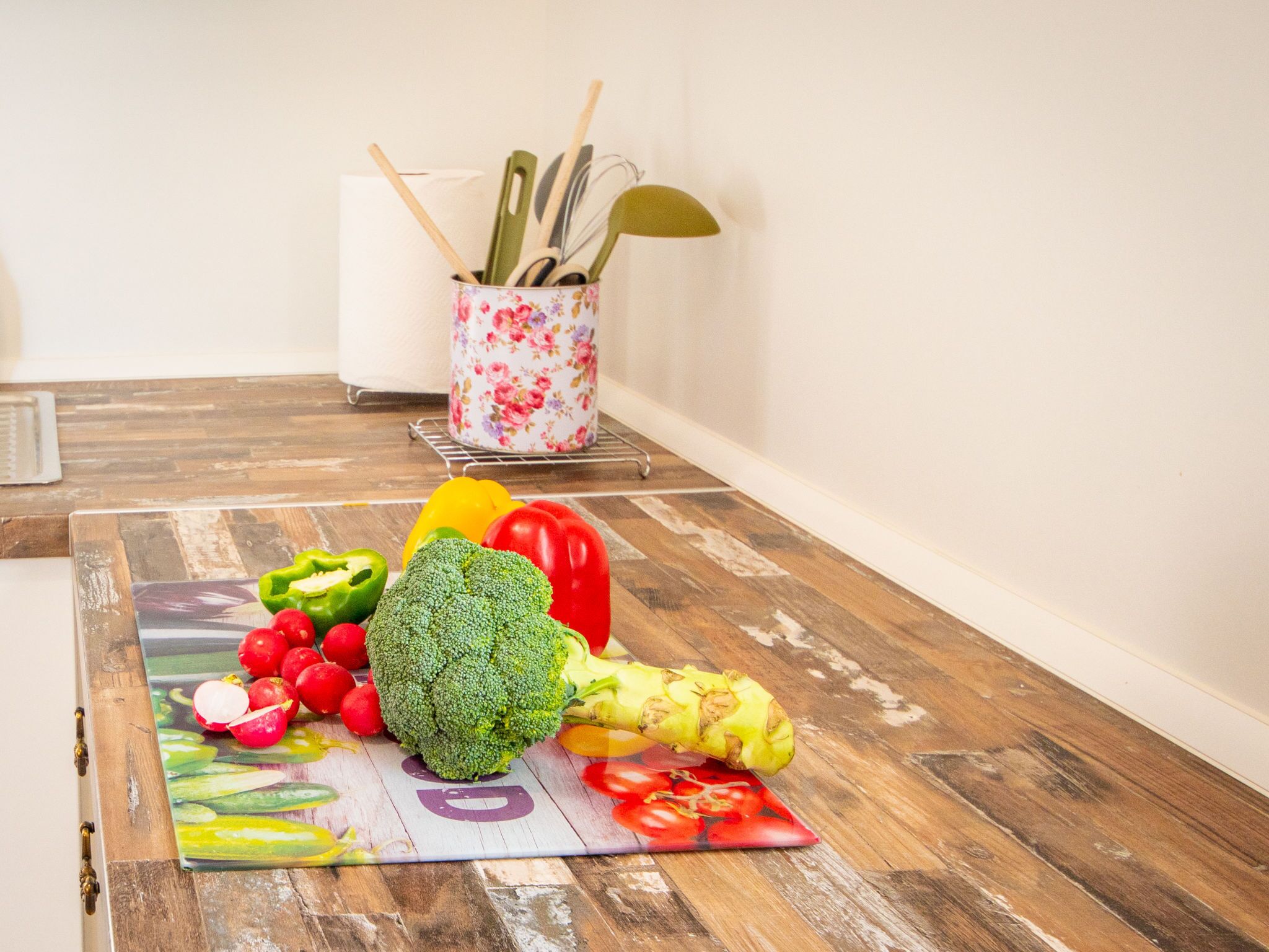 Un plan de travail de cuisine avec une planche à découper sur laquelle sont disposés divers légumes, ainsi qu’un récipient décoré de motifs floraux contenant des ustensiles de cuisine.