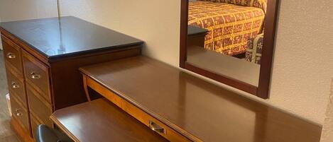 A hotel room with a wooden desk, a chair, a flat-screen TV, a bedside table, and a large mirror reflecting the bed and a painting on the wall.