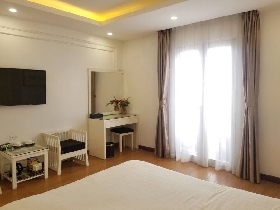 A hotel room with a large bed, a desk with a chair, a TV mounted on the wall, and a window with curtains.