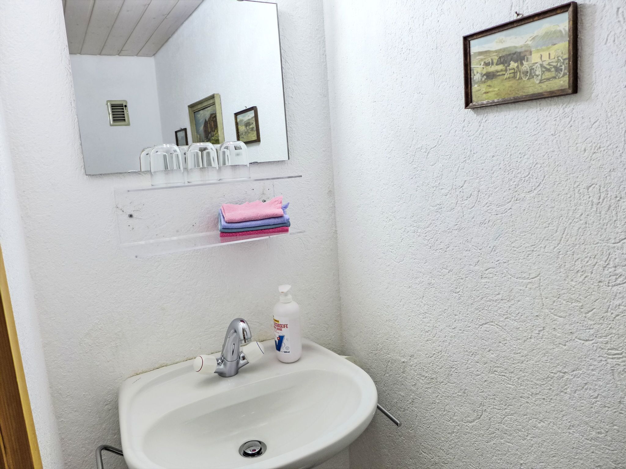 Un baño con un lavabo blanco, un espejo, un estante con toallas dobladas y un cuadro en la pared.