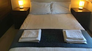 Un lit bien fait, avec du linge de lit blanc, deux oreillers et deux serviettes pliées, posés sur un couvre-lit de couleur foncée. De chaque côté du lit, on trouve une table de chevet avec une lampe.