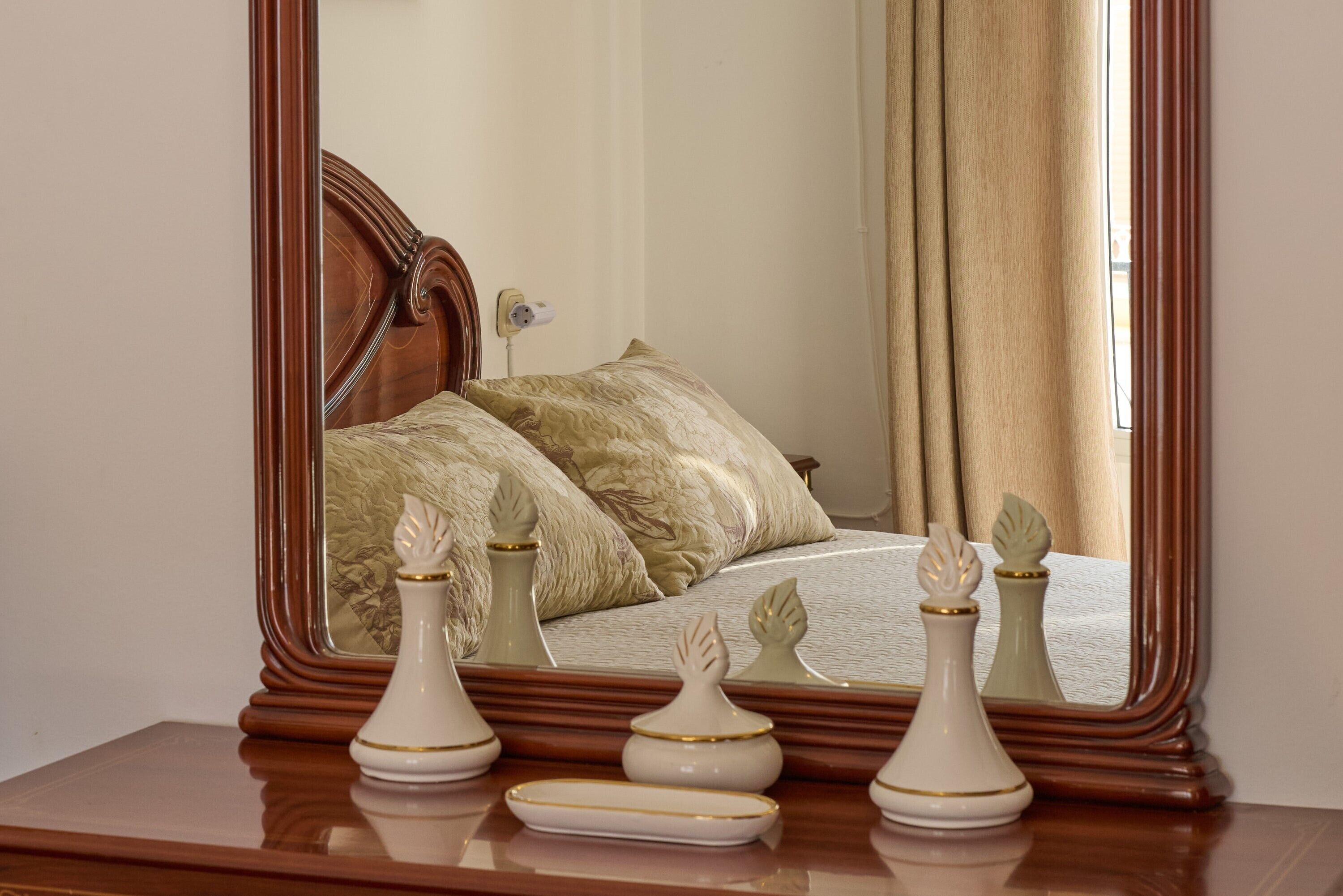 A bedroom with a large mirror, a wooden headboard, a bed with beige bedding, and decorative vases on a dresser.