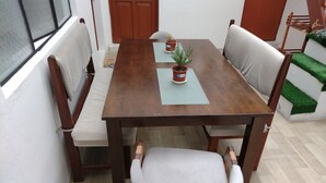 Un coin repas avec une table en bois, quatre chaises, deux plantes en pot et une fenêtre avec des stores.