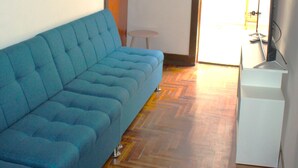 Une pièce avec un canapé bleu, une petite table blanche et un téléviseur. Il y a une porte qui donne sur une autre pièce et une fenêtre avec vue.