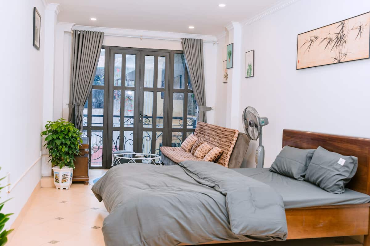 Un dormitorio con cama, mesita de noche, planta en maceta, ventilador y vista al balcón exterior.