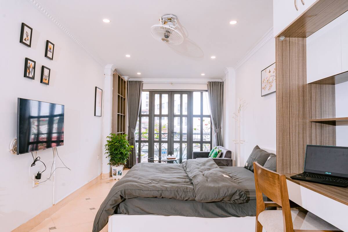 Un dormitorio moderno con una cama, un escritorio con una computadora portátil, un televisor y ventanas amplias con cortinas.