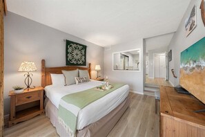 Ein Schlafzimmer mit einem großen Bett, zwei Nachttischen, einem kleinen Tisch, einer grünen Tagesdecke, einem Zierkissen, einem Wandbild, einem Spiegel und einem Fernseher auf einem Holzständer.