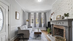 Ett vardagsrum med en grå soffa, ett soffbord, en öppen spis och ett stort fönster med gardiner.