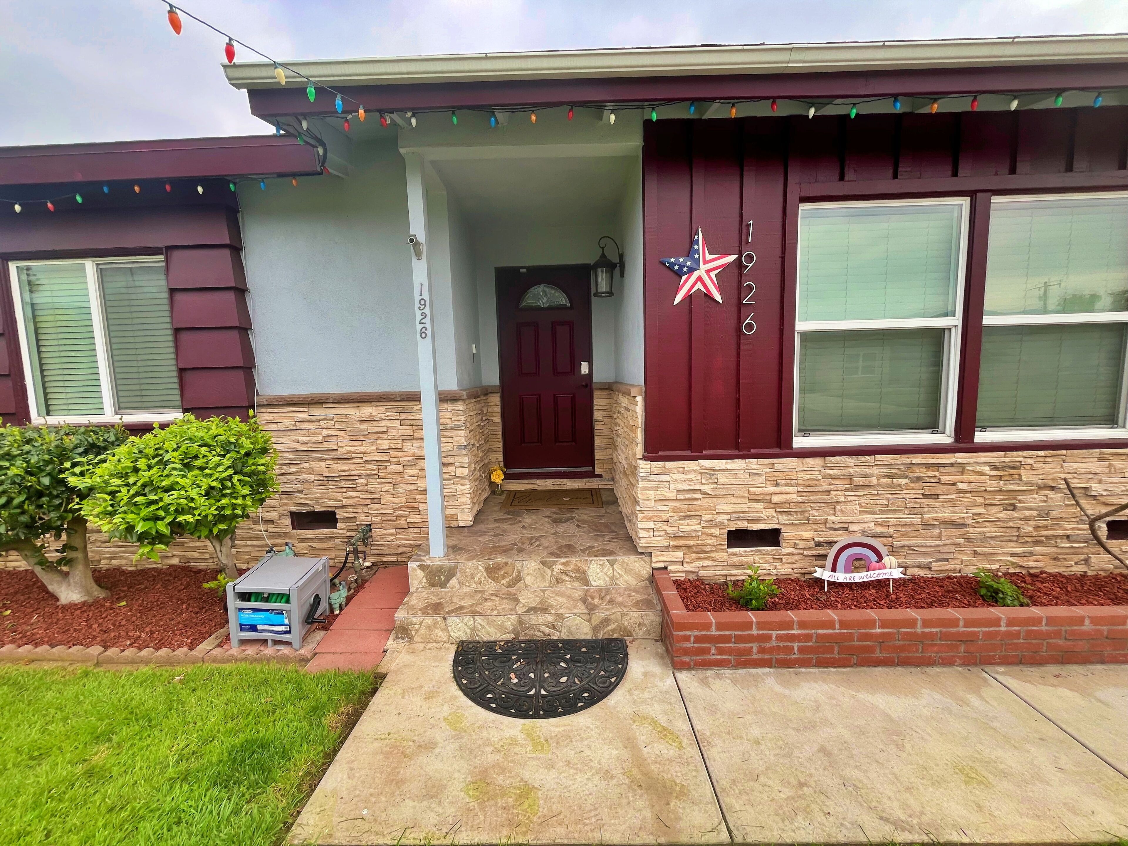 Uma casa com porta da frente, caixa de correio e um pequeno jardim com um banco e uma placa que diz 'ALL ARE WELCOME'.
