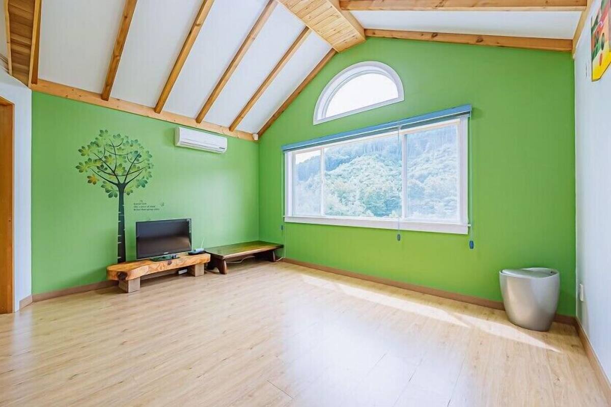 Una habitación con paredes verdes, piso de madera, un ventanal con vistas a árboles, un televisor pequeño sobre un mueble de madera y un cubo de basura.