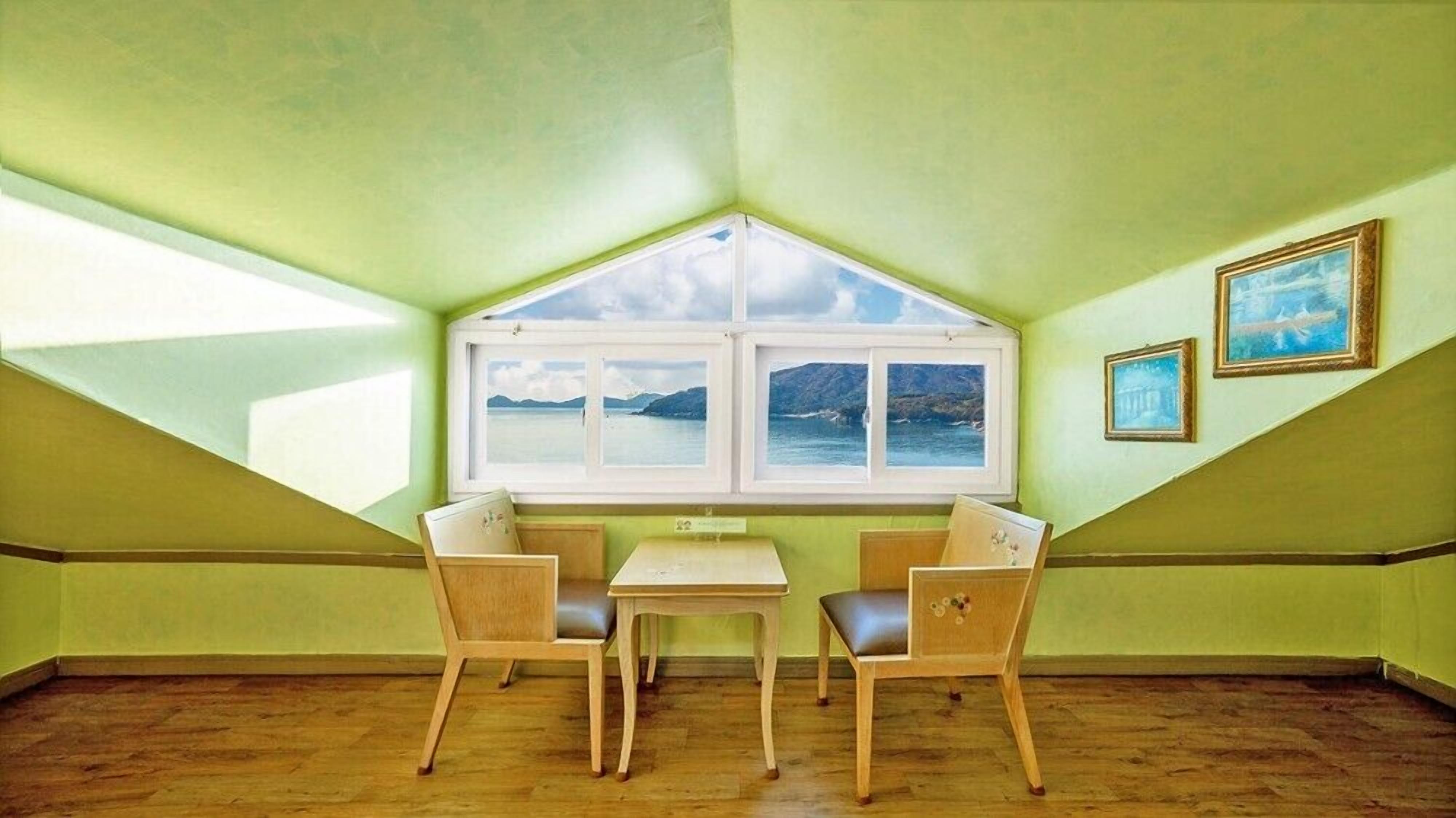 A room with a sloped ceiling, a bay window with three panes, two wooden chairs, a small table, and two framed pictures on the wall.