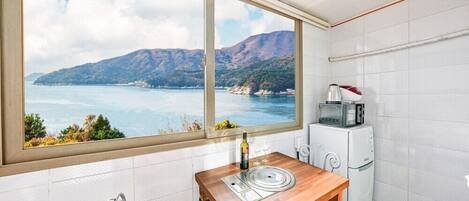 A kitchen with a view of a coastal landscape, a small table with a bottle and glasses, and a microwave on a white cabinet.