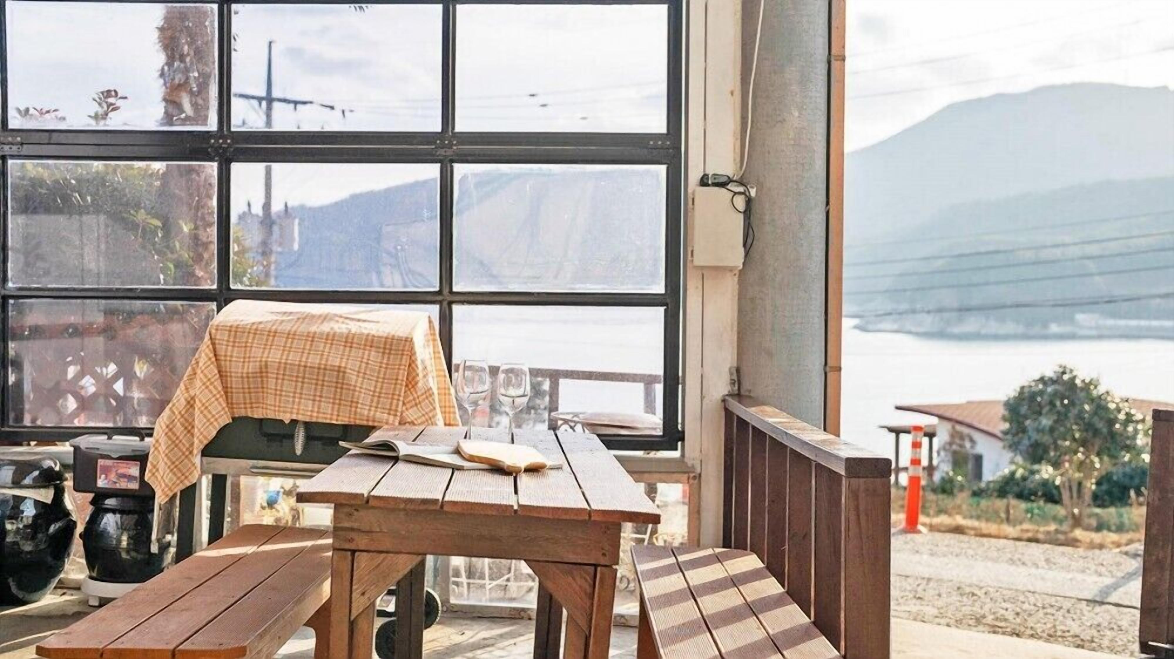 A wooden table with a checkered tablecloth, a bench, and a view of a body of water and mountains through large windows.