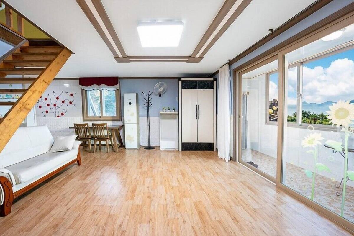 Un amplio espacio habitable con una escalera de madera, un sofá blanco, una mesa de comedor con sillas, un refrigerador y un gran ventanal corredero que da a un balcón con vista al mar.