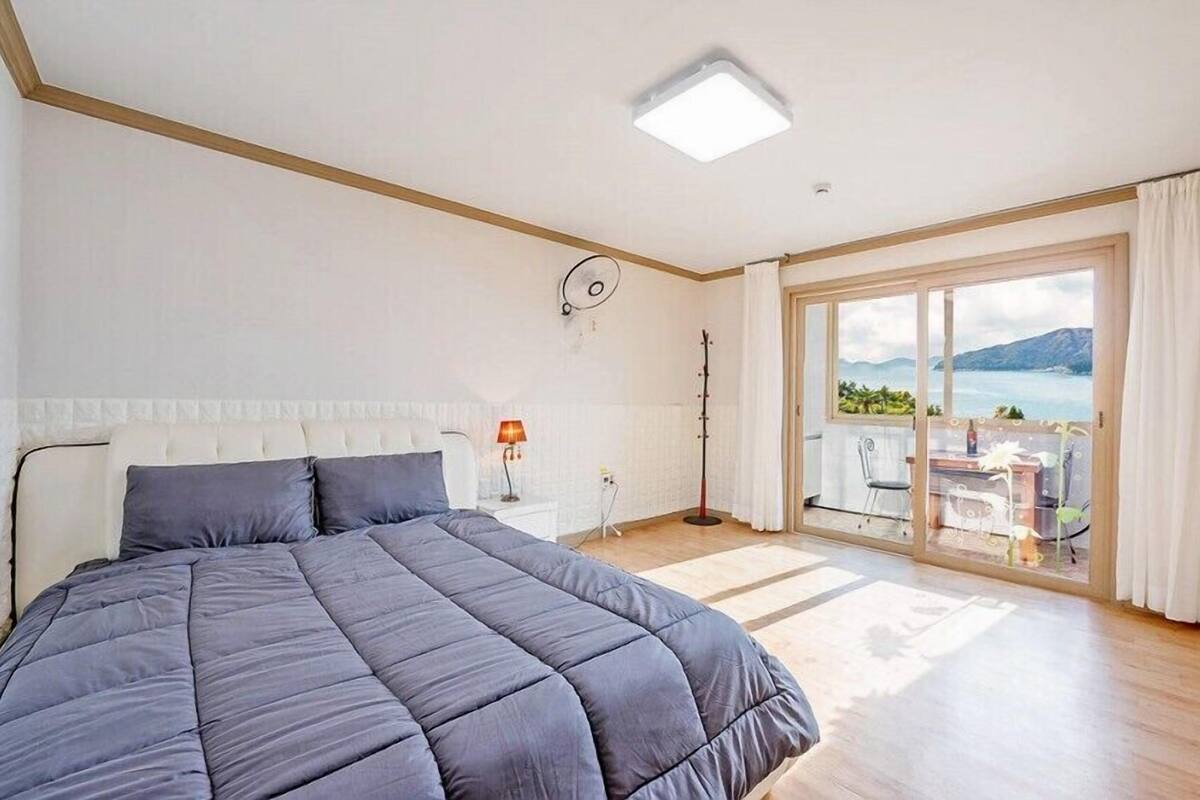 Un dormitorio con una cama grande, un escritorio, una silla, una lámpara y vista al mar a través de una puerta corrediza de vidrio.