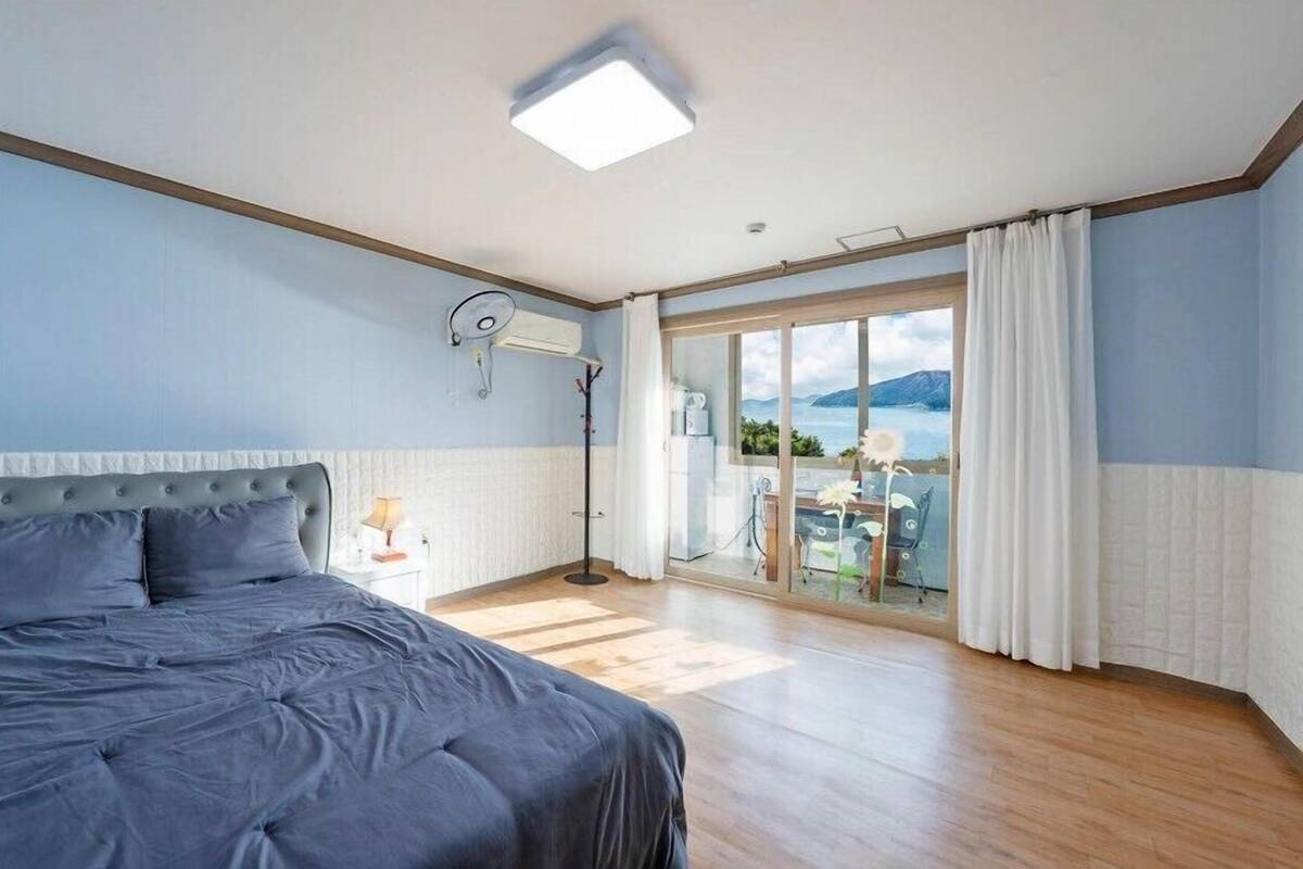 Un dormitorio con una cama grande, una lámpara en el techo, un equipo de aire acondicionado, vistas al exterior a través de un ventanal y un balcón con mesa y sillas.