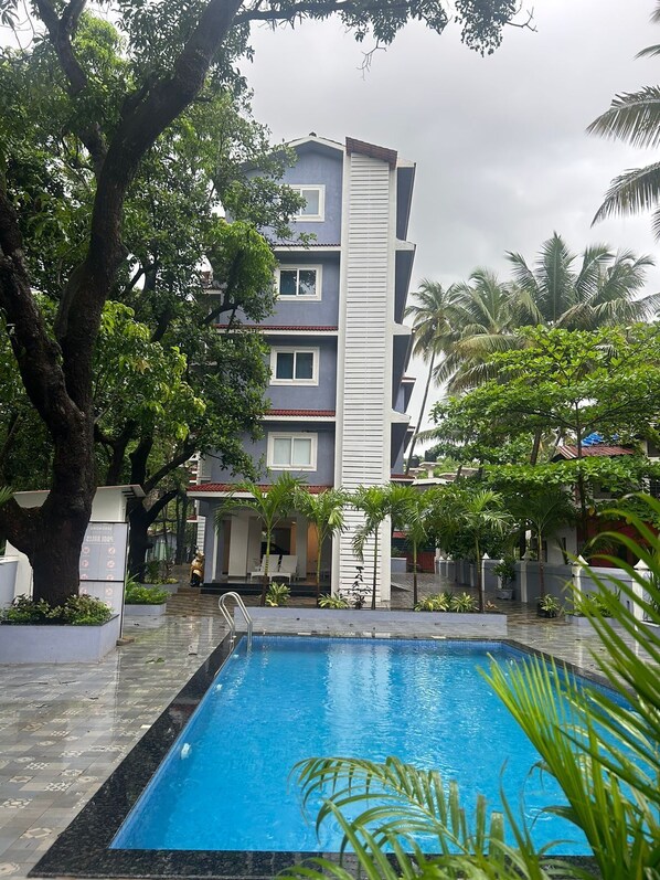 Una piscina con borde de baldosas, rodeada de exuberante vegetación y un edificio de varios pisos con balcones.