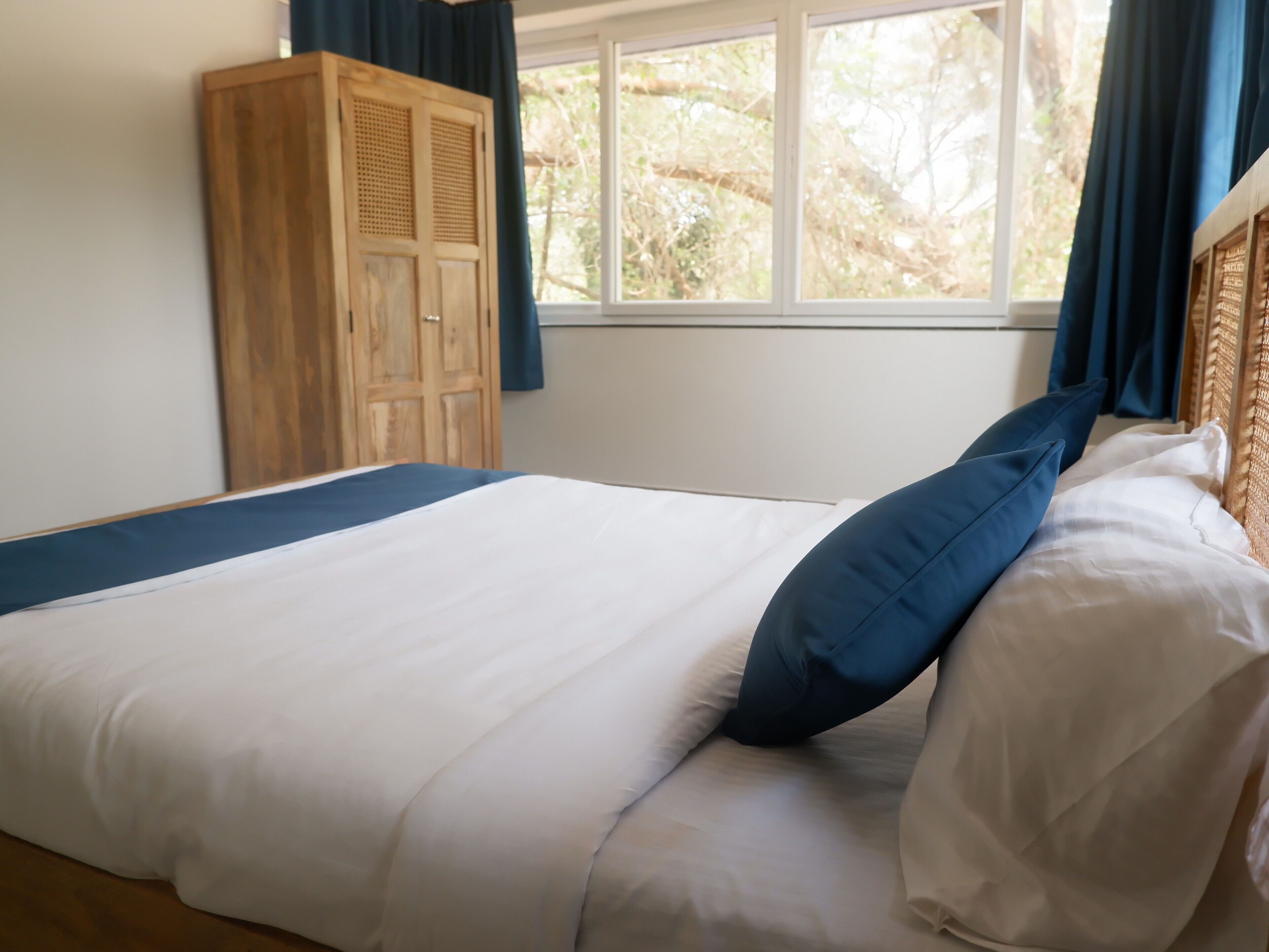Una cama bien hecha con ropa de cama blanca y azul, un armario de madera y una ventana con cortinas.
