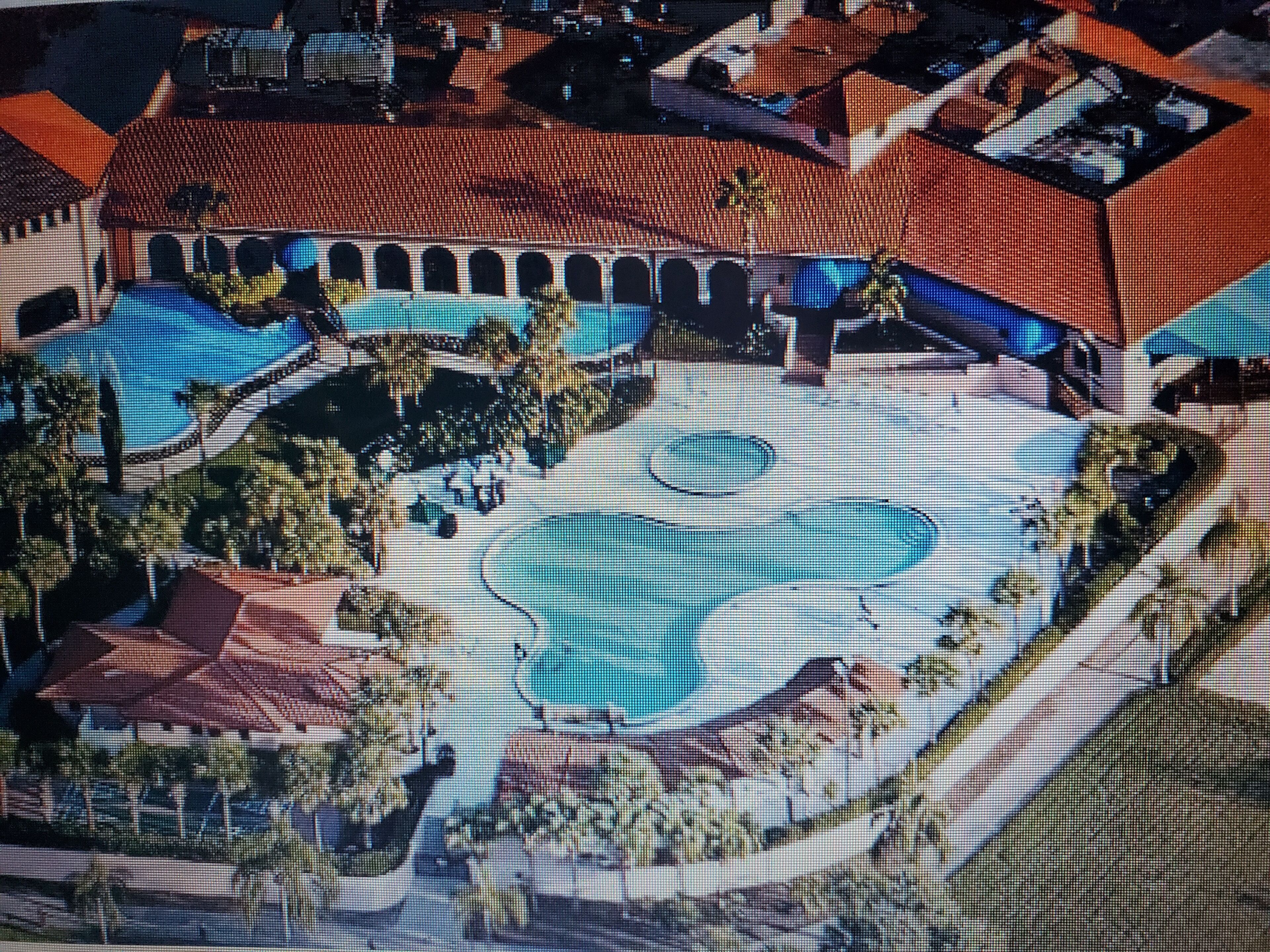 Vue aérienne d’un complexe hôtelier doté de plusieurs piscines, d’un bâtiment central avec un toit rouge et d’espaces paysagers environnants.