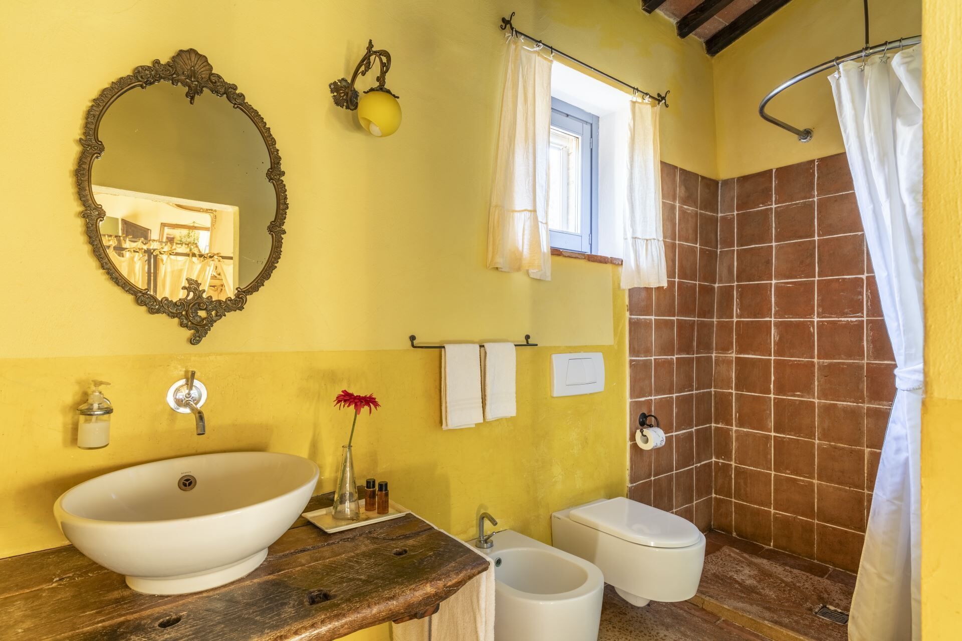 Un baño con un lavamanos ovalado grande, un inodoro, una ducha con cortina, un espejo con un marco ornamentado y una ventana con cortina.