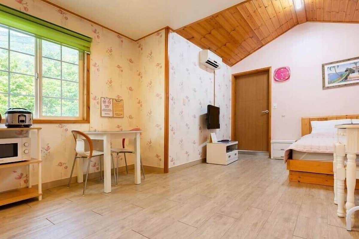 A room with a wooden ceiling, a bed, a desk with a chair, a window with a green blind, and a door.