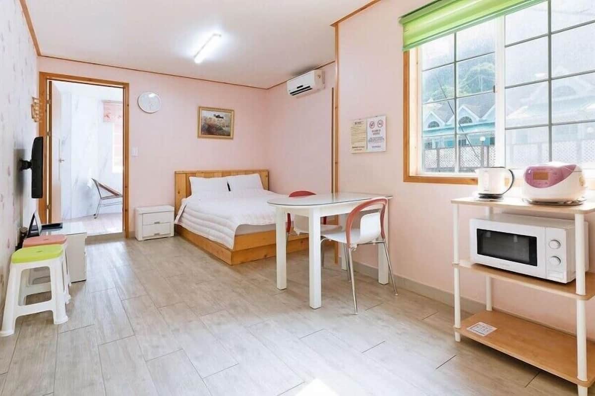 A small, well-lit room with a bed, a table, a chair, a microwave, and a window with blinds.