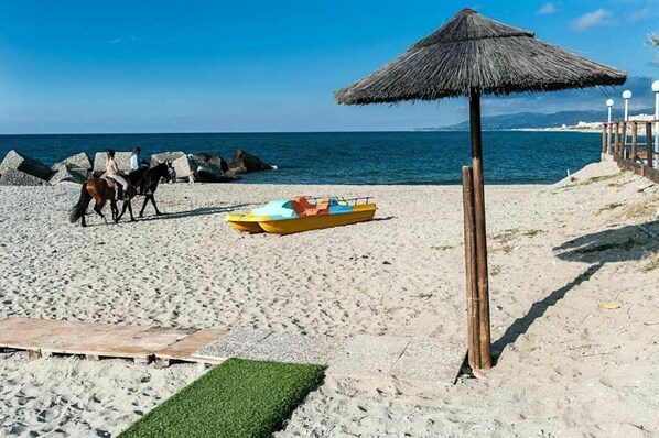 A beach with a thatched umbrella, a horse, and a small boat.
