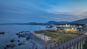 Un edificio moderno frente al mar con fachada de vidrio, ubicado en una costa rocosa con un camino pavimentado que lo lleva a él.