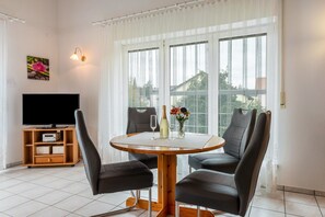 Un coin repas comprenant une table ronde, quatre chaises, un téléviseur posé sur un meuble et une fenêtre avec des rideaux.