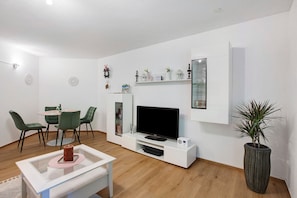 A modern living room with a flat-screen TV, green chairs, a white coffee table, and a potted plant.