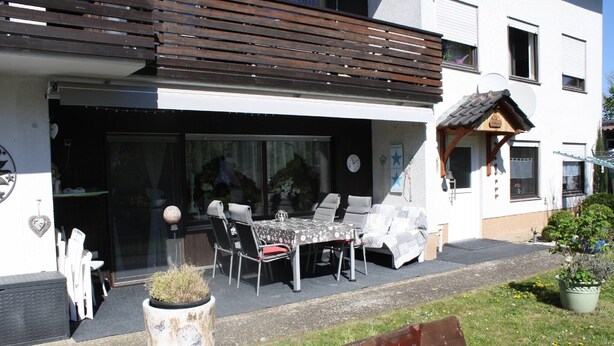 A modern building with a patio area featuring a table and chairs, a potted plant, and a wooden bench.