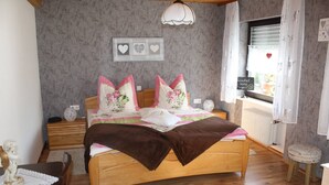 A bedroom with a wooden bed, a nightstand, a bedside table, a window with curtains, and a wall with heart-shaped decorations.