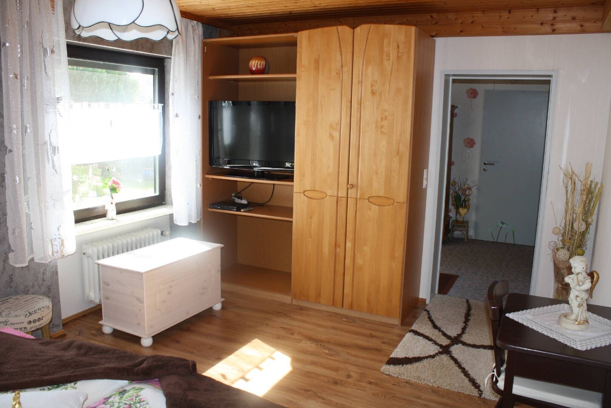 Ett rum med en garderob i trä, en tv, en vit byrå, ett fönster med gardiner och ett litet bord med en dekorativ prydnad.