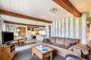 A living room with a television, wooden coffee table, and a sofa. There is a kitchen in the background with white cabinets and a dining area.