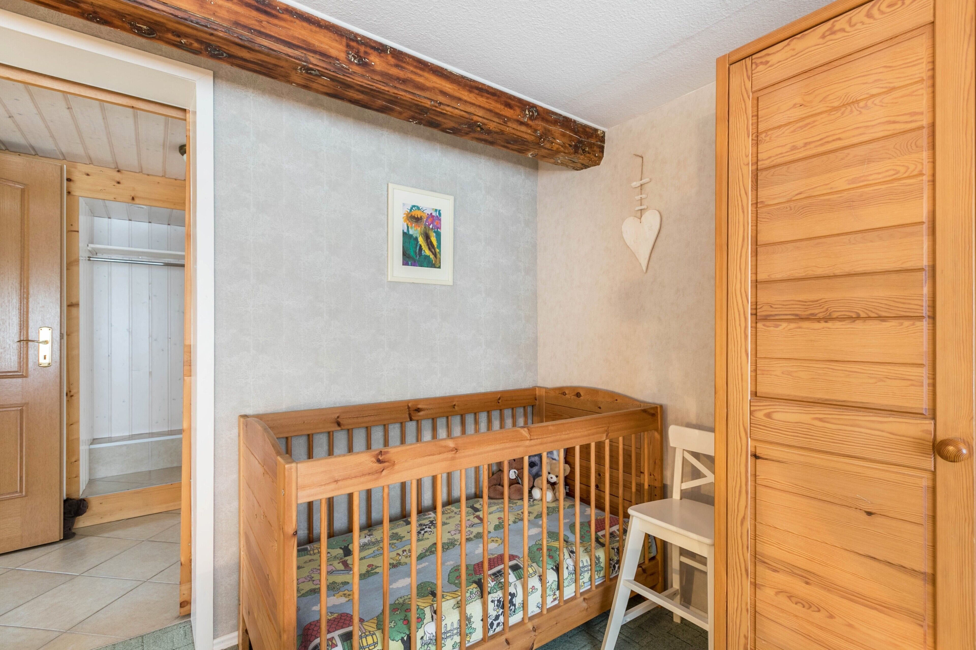 Ein hölzernes Kinderbett mit einem gemusterten Lattenrost, ein weißer Stuhl und ein Holzschrank in einem Raum mit schräg verlaufender Decke.