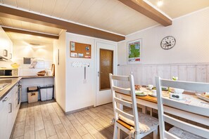 Una cocina con microondas, lavamanos y armarios de madera. Hay una mesa de comedor con sillas y un reloj en la pared.