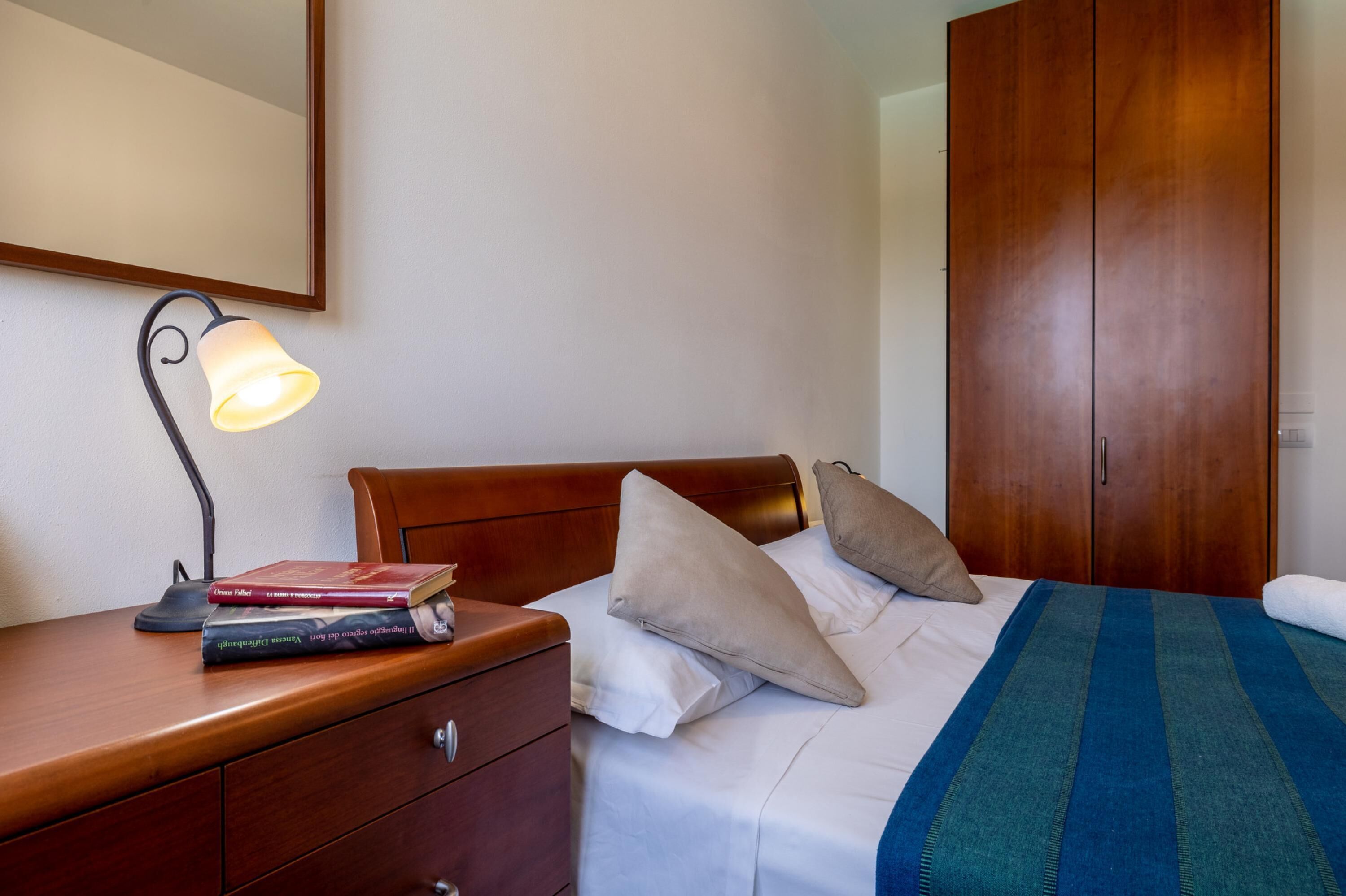 A neatly made bed with pillows, a bedside lamp, a wooden nightstand with books, and a wardrobe.