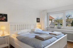 A white bed with two pillows, a bedside table with a lamp, and a window with a view of trees and buildings.