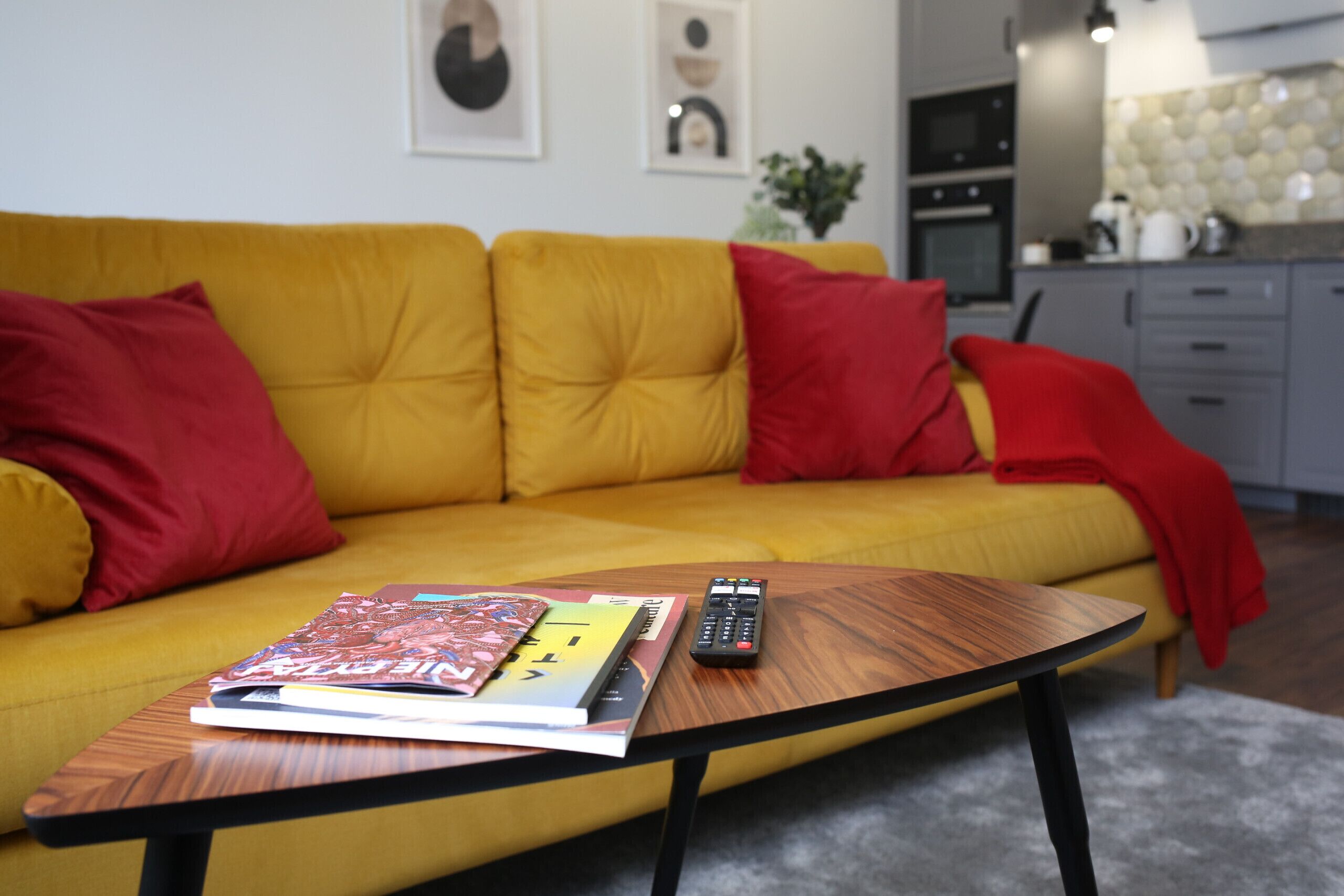 Un canapé jaune avec des coussins rouges, une table basse avec une télécommande et un magazine, et une cuisine en arrière-plan.