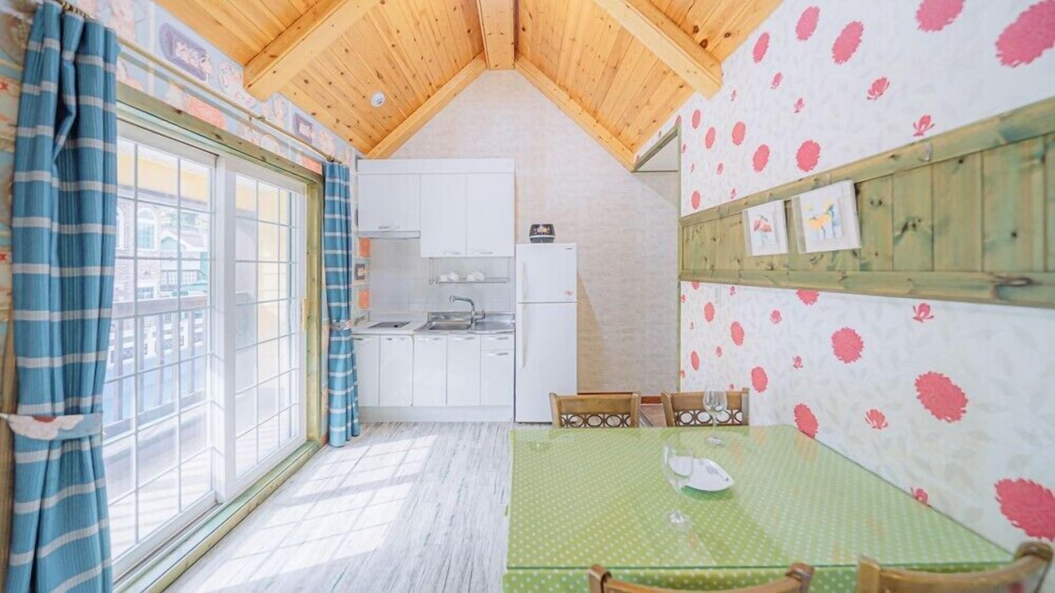 A compact kitchen with white cabinets, a sink, and a refrigerator. There's a dining table with chairs and a window with blue curtains.
