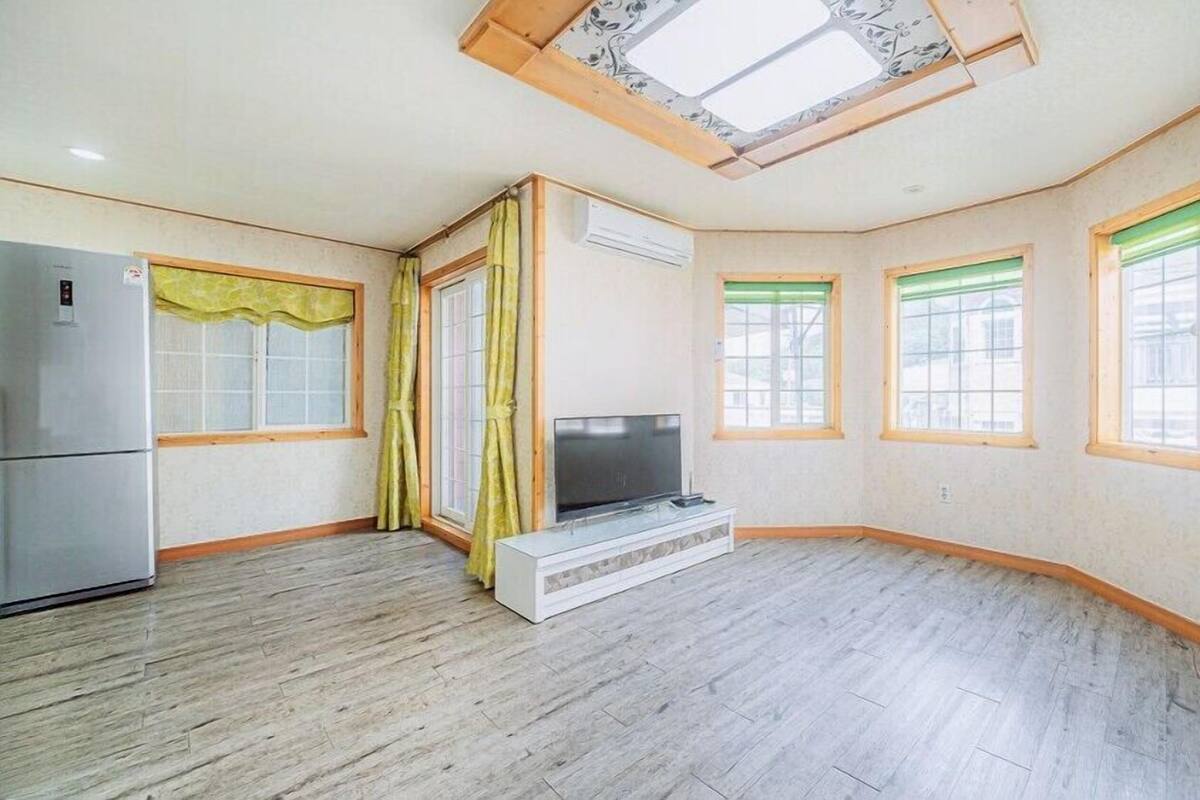 Amplia sala de estar con televisor de pantalla plana, piso de madera y ventanas grandes con persianas verdes.