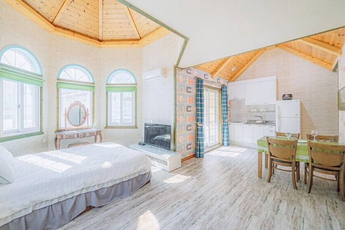 Un dormitorio con una cama grande, un tocador, un espejo y una televisión. Incluye una kitchenette con refrigerador, lavamanos y estufa. La habitación tiene techo inclinado y ventanas amplias.