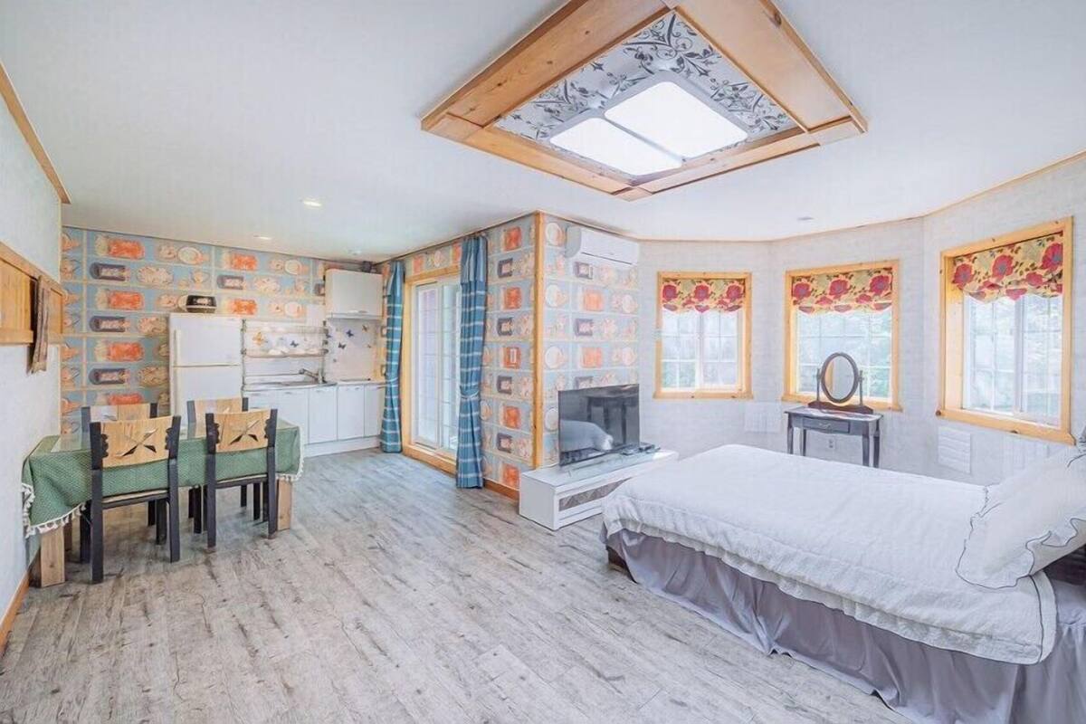 Un dormitorio con cama, televisor, escritorio con silla, ventana con cortinas y tragaluz.