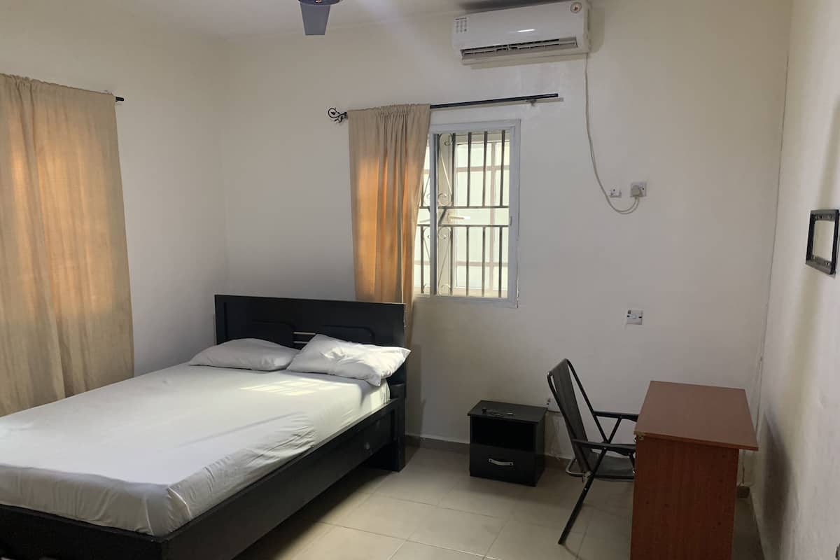 A bedroom with a bed, a desk, a chair, an air conditioner, and a window with curtains.