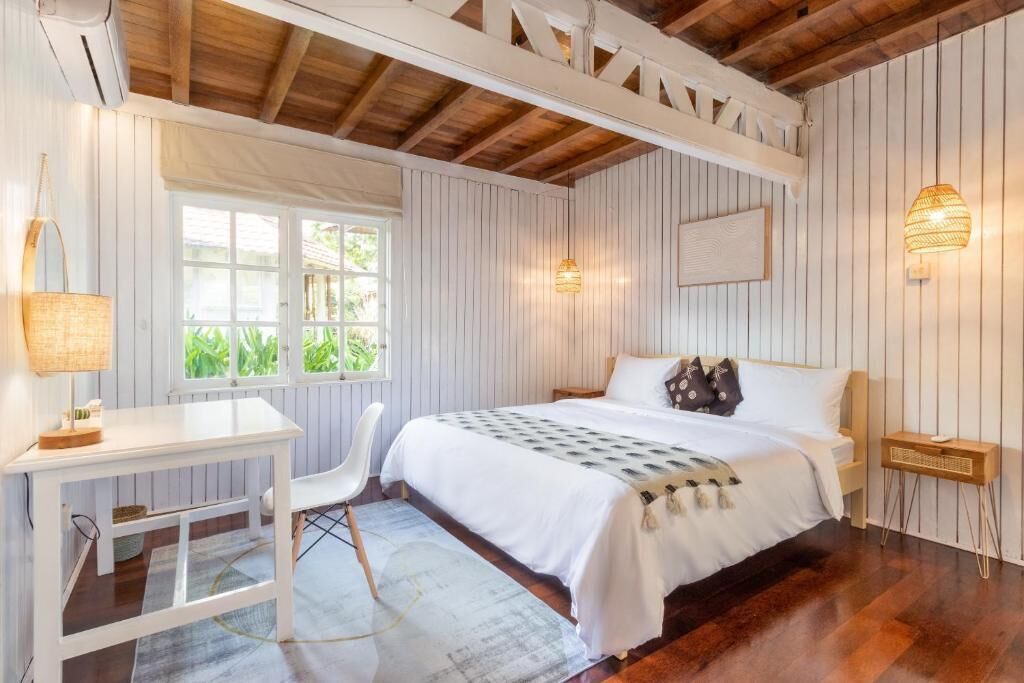 A bedroom with a bed, a desk, and a chair. There are windows and a ceiling with wooden beams.
