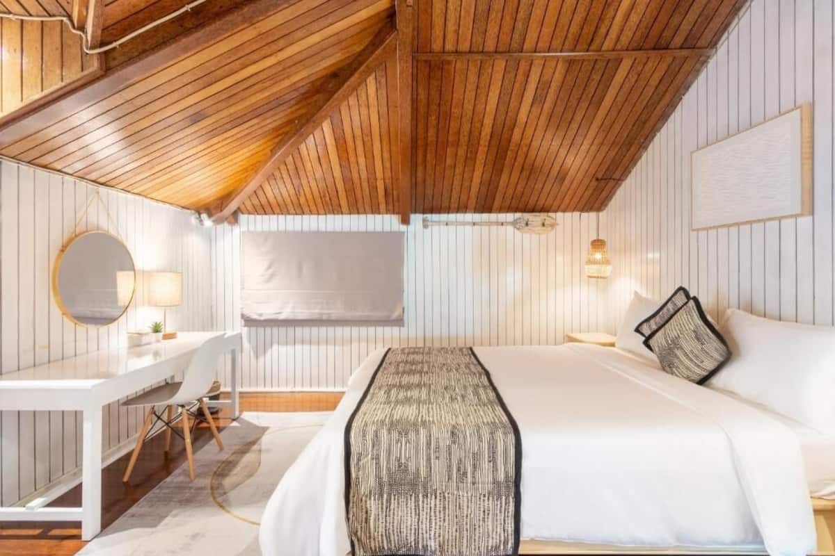 Quarto com teto de madeira, cama branca, escrivaninha com cadeira, espelho redondo e luminária de cabeceira.