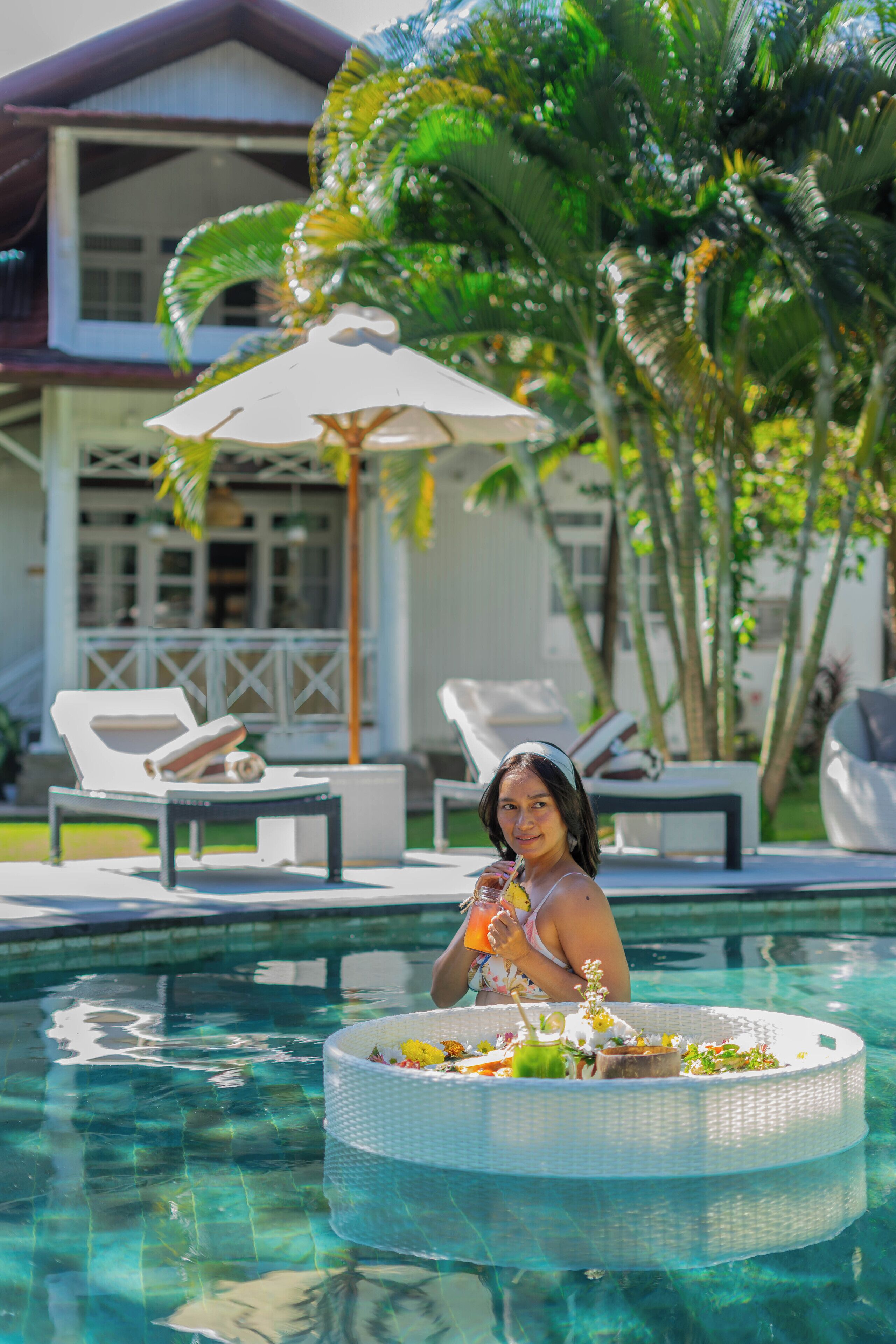 A pool area with lounge chairs and umbrellas, a floating platform with flowers and drinks, and a house in the background.